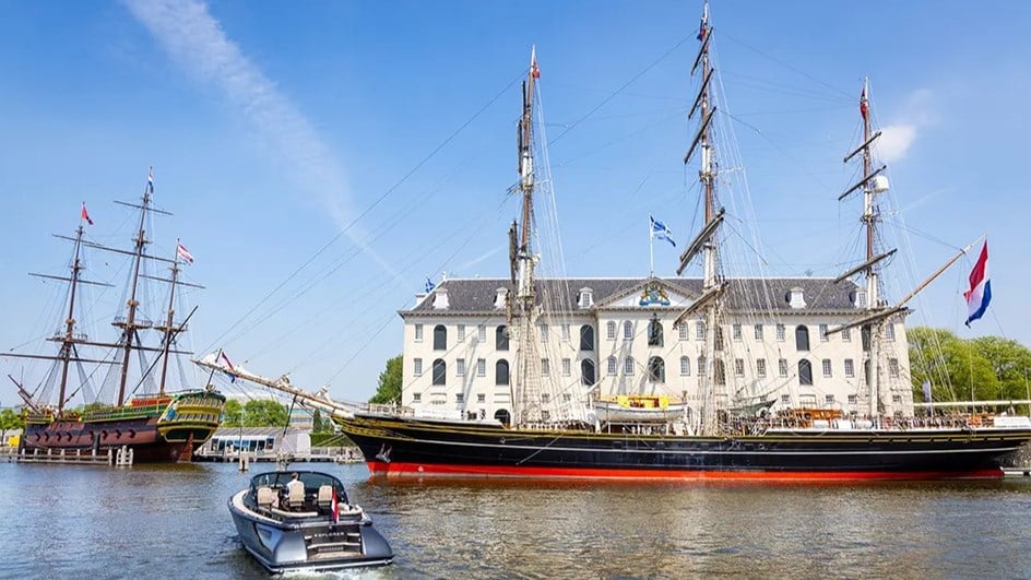 A boat sailing near a number of old vessel ships in front of a manor